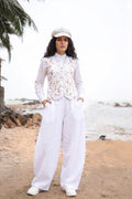 A woman stands on a sandy beach wearing a white outfit consisting of a long-sleeve top with a floral embroidered vest, wide-leg pants, and a white cap.