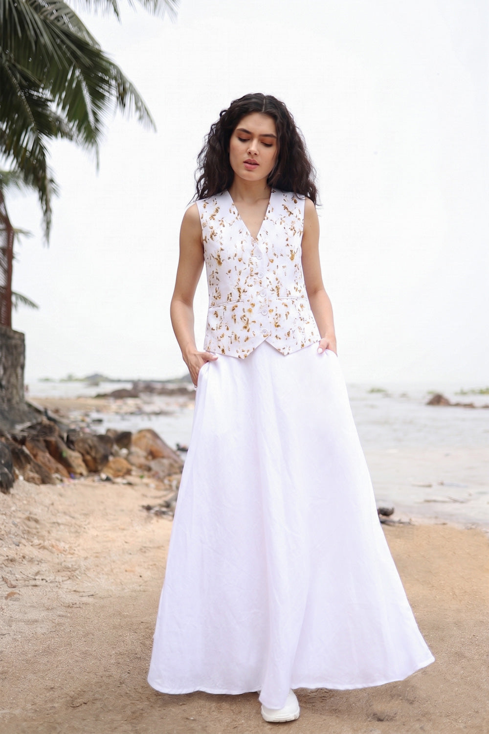 A woman wearing a sleeveless white embroidered vest and a long white skirt with pockets, standing on a sandy beach with water and palm trees in the background.