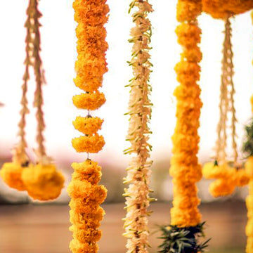 Decorative hanging flower garlands, including bright orange marigold strands and white floral garlands, suspended against a softly blurred light background.