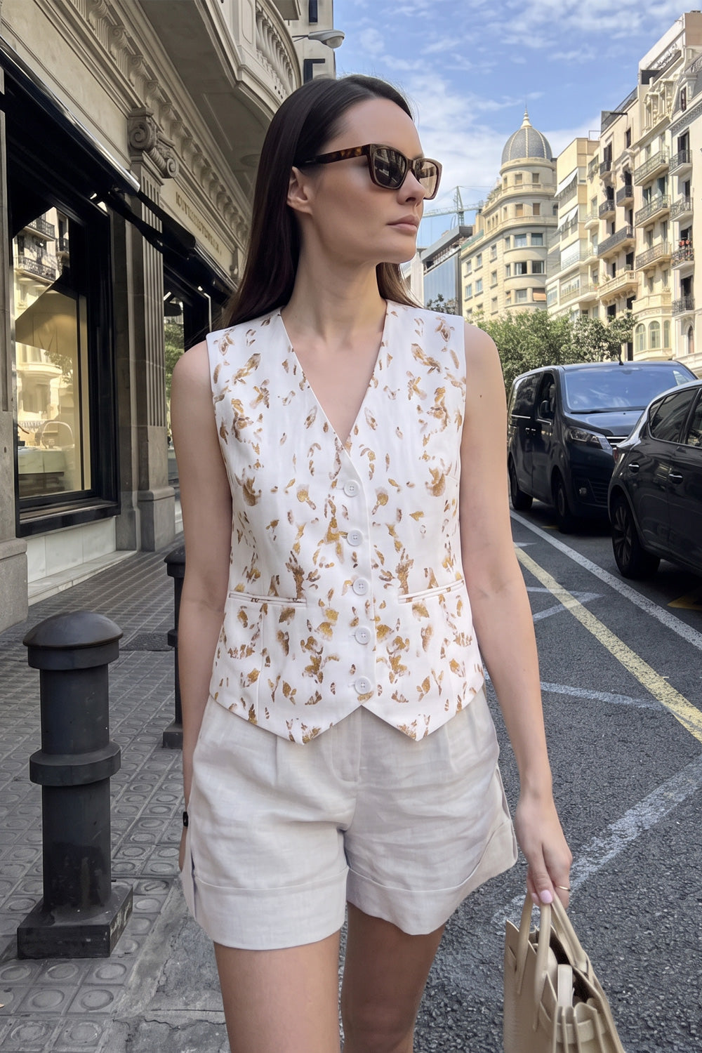 A woman stands on a city street wearing a sleeveless white wrap-style vest with a tan floral botanical-print and light beige shorts, holding a beige handbag.
