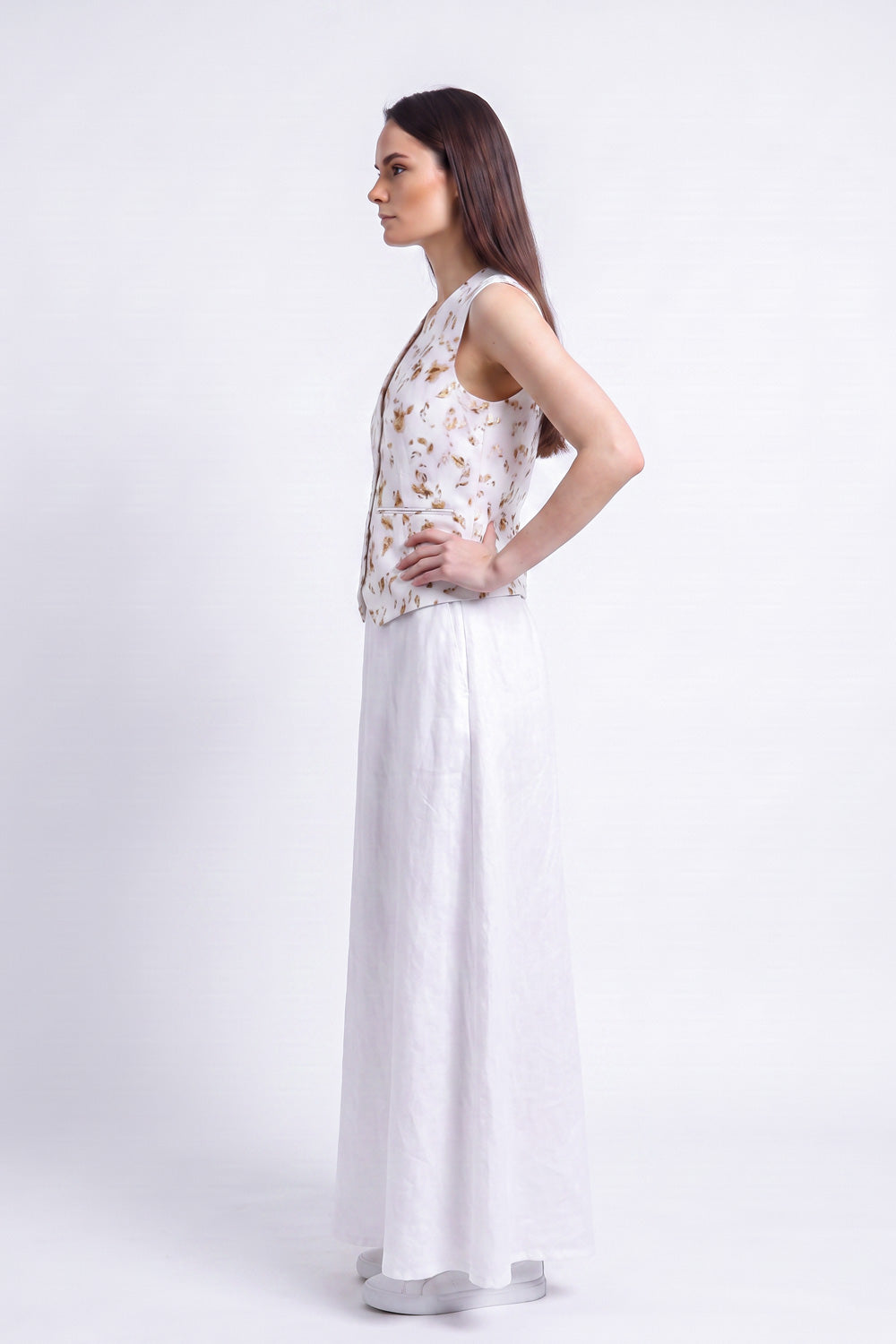 A woman stands in profile wearing a sleeveless white top with a small beige and brown floral eco-print, paired with a long white skirt, against a plain light gray studio background.