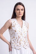 A woman modeling a sleeveless white vest with a gold abstract botanical-print, paired with a white skirt, against a light gray studio background.