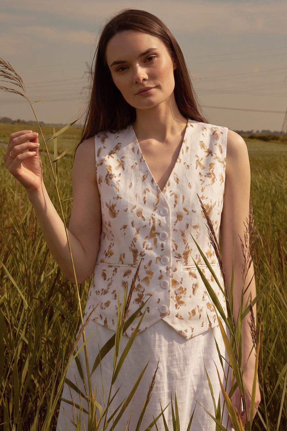 A woman standing in a grassy field wearing a sleeveless white vest with a brown floral botanical and a white skirt.