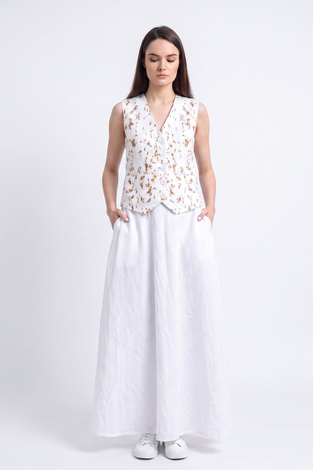 A woman stands against a light gray studio background wearing a sleeveless white vest with a gold floral eco-print and a long white skirt.