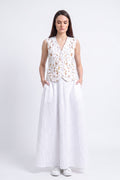 A woman stands against a light gray studio background wearing a sleeveless white vest with a gold floral eco-print and a long white skirt.