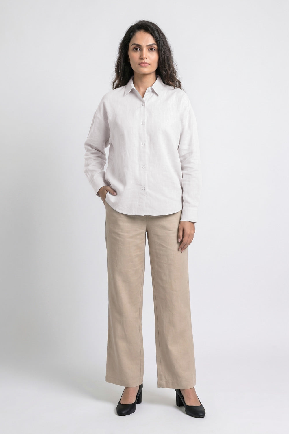 A woman modeling a white long-sleeve linen button-up shirt and beige wide-leg linen trousers against a light gray studio background.