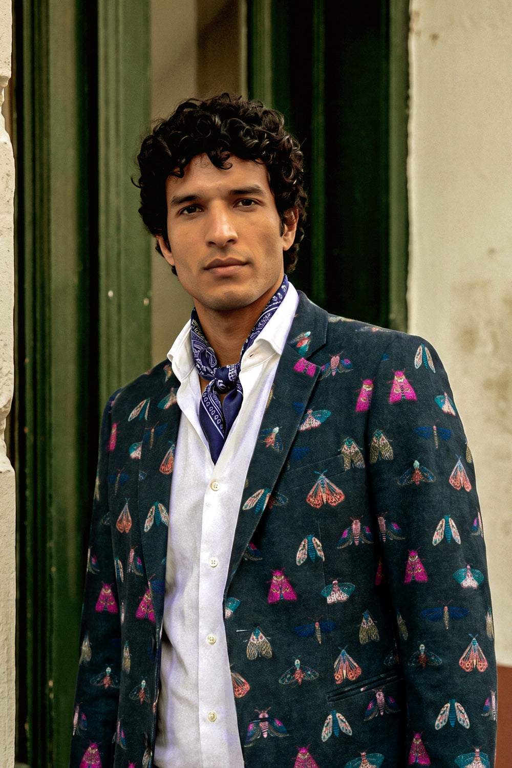 A man wearing a dark teal blazer covered in colorful moth and butterfly patterns, styled with a white shirt and a blue patterned necktie, standing in front of a rustic green and beige doorway.