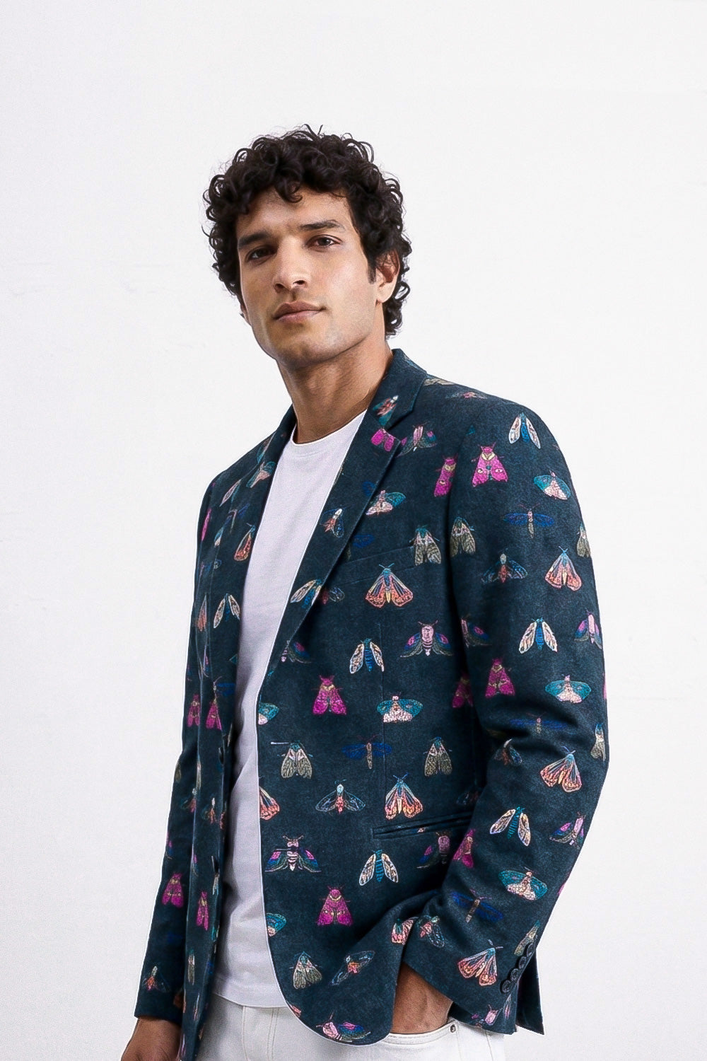 A man wearing a dark teal blazer with a colorful butterfly and moth pattern over a white t-shirt, photographed against a plain white background.