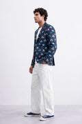 A man wearing a navy blue patterned blazer, white trousers, and navy blue canvas sneakers, standing in profile against a light gray studio background.