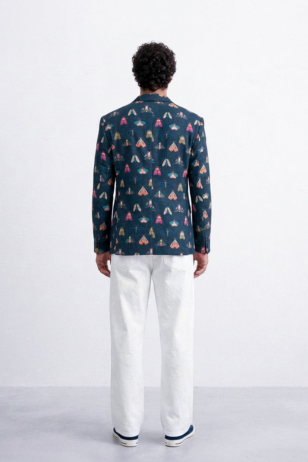 A man viewed from behind wearing a navy blue blazer with a colorful pattern of small tents and houses, paired with white trousers and navy sneakers, against a plain white studio background.