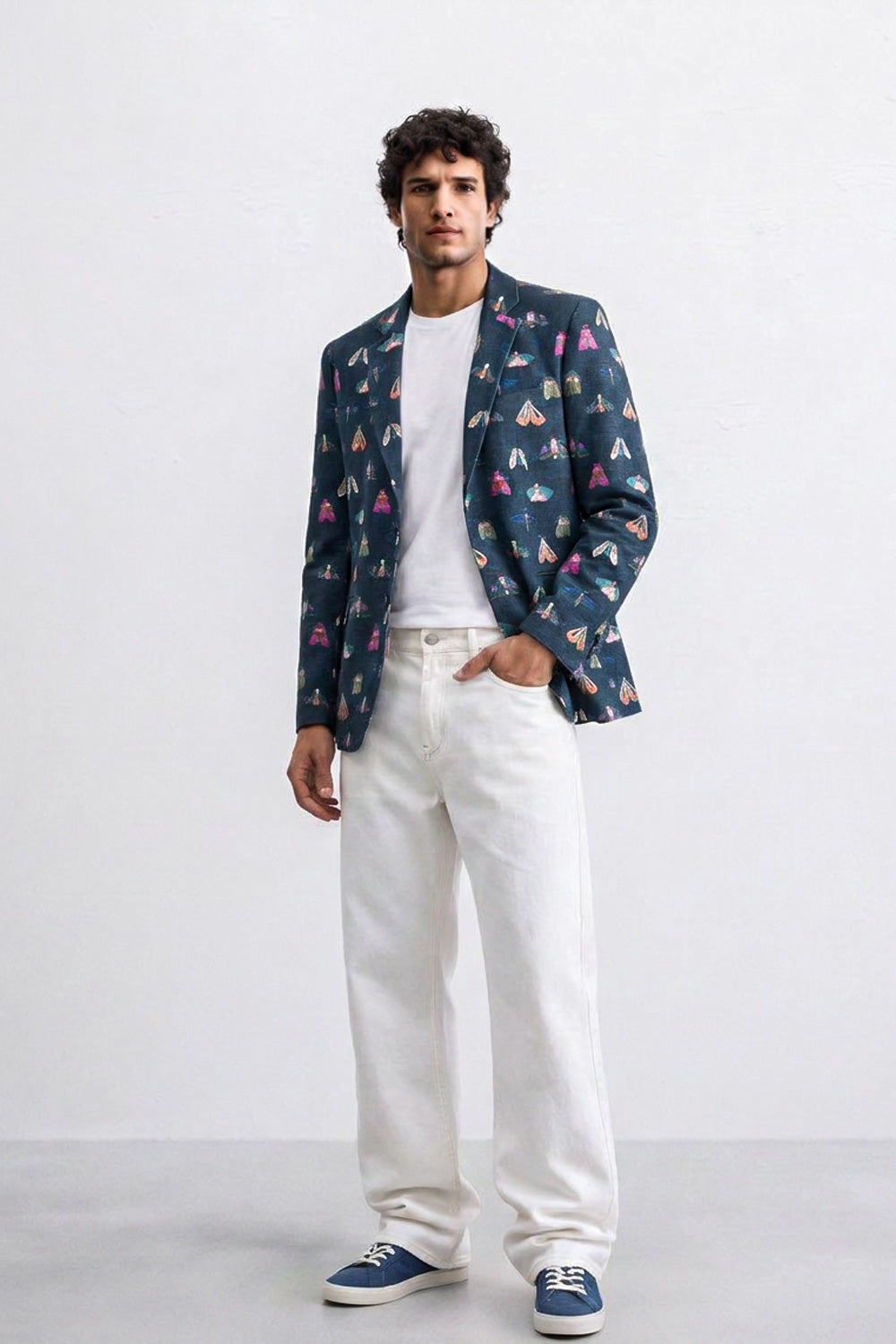 A man modeling a navy blue blazer with a colorful geometric pattern, worn over a white t-shirt and white jeans, paired with blue sneakers, against a plain white studio background.
