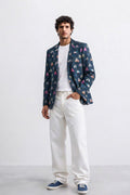 A man modeling a navy blue blazer with a colorful geometric pattern, worn over a white t-shirt and white jeans, paired with blue sneakers, against a plain white studio background.