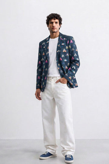 A man modeling a navy blue blazer with a colorful geometric pattern, worn over a white t-shirt and white jeans, paired with blue sneakers, against a plain white studio background.
