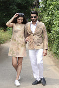 A couple walking arm-in-arm on a paved path surrounded by lush green foliage. The woman wears a beige sleeveless dress with a green leaf eco-printed  pattern and white sneakers.