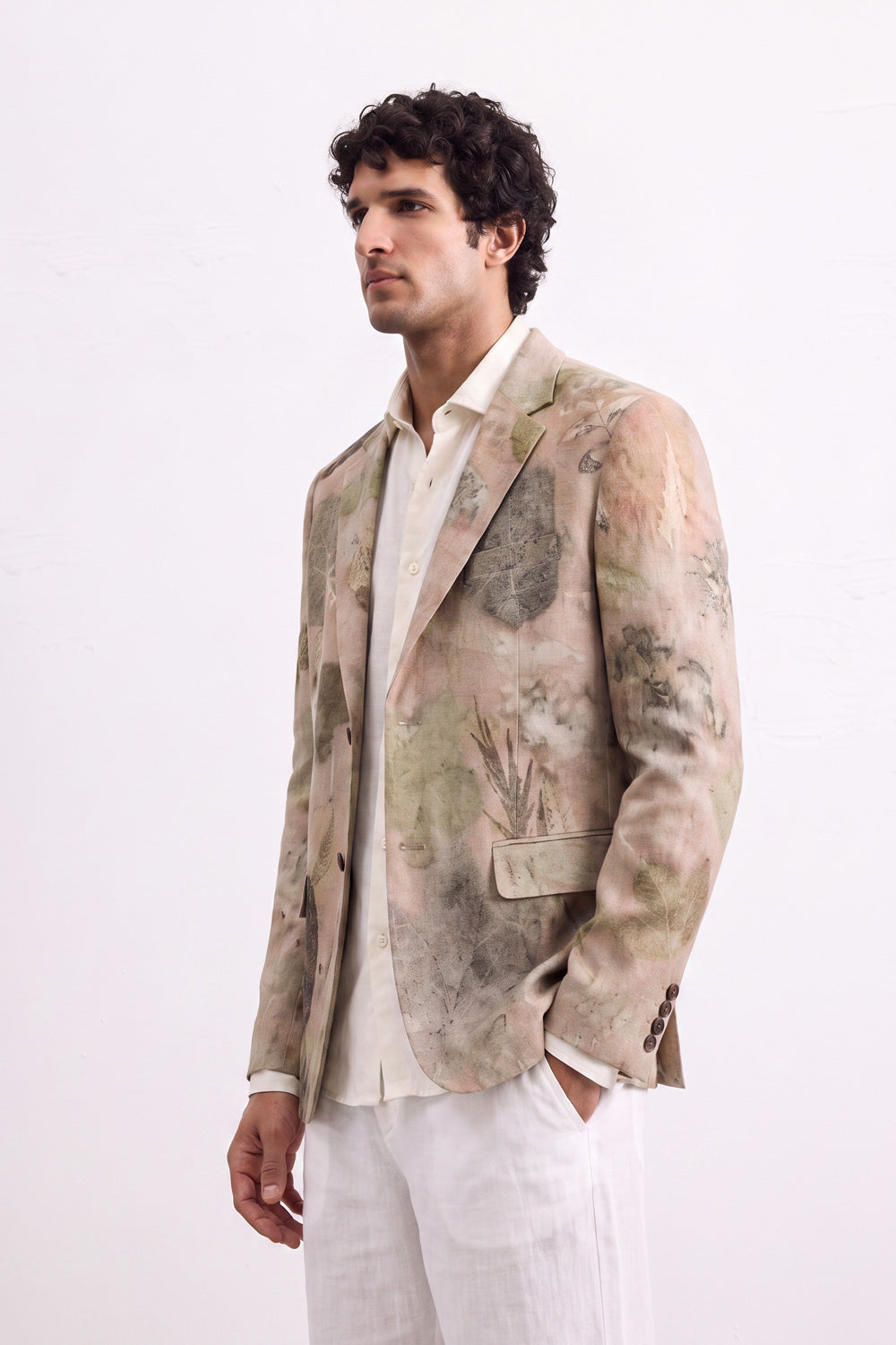 A man wearing a light beige and green eco-painterly patterned blazer over a cream shirt and white trousers, standing against a plain white studio background.