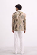 A man viewed from the back wearing a light beige blazer with a muted green and gray botanical leaf pattern, paired with white trousers, against a plain white studio background.