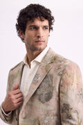 A man with curly dark hair wearing a light beige linen-blend blazer with a botanical leaf eco-print and a white collared shirt, posed against a plain white studio background.