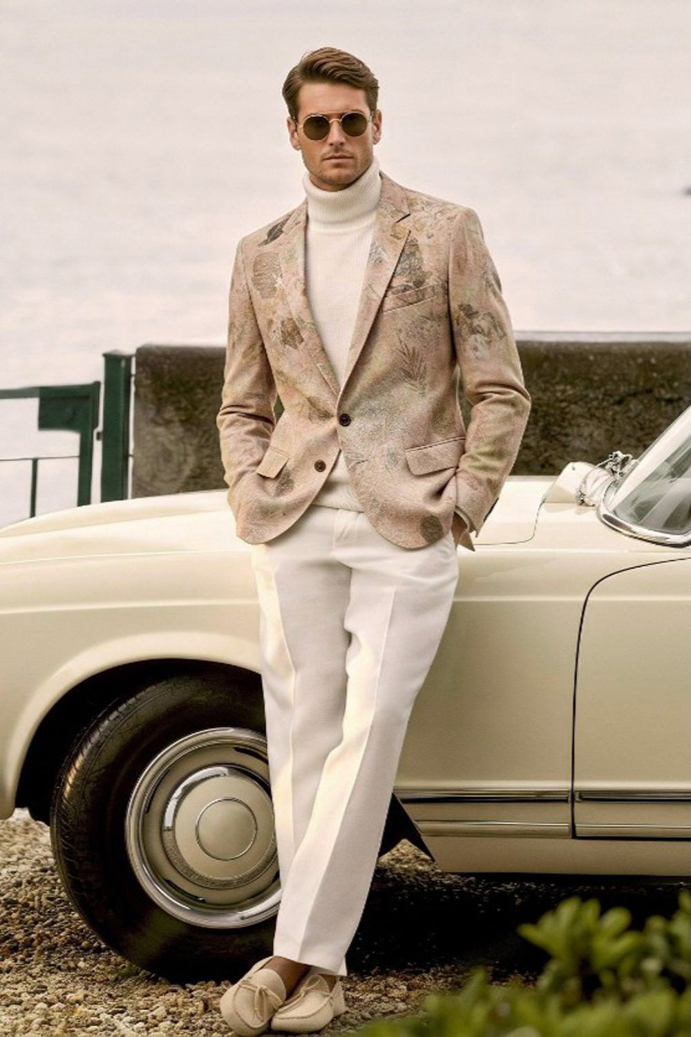 A man wearing a beige eco-printed patterned blazer, white turtleneck, and white trousers leans against a cream-colored vintage car outdoors.