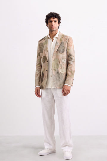 A man modeling a light beige and white abstract-patterned eco-print blazer over a white shirt and white trousers, standing against a plain white studio background.