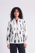 A model with curly dark hair wearing a white zip-up hemp bomber jacket with a black eco-print, styled over a red shirt and black pants, against a light gray studio background.