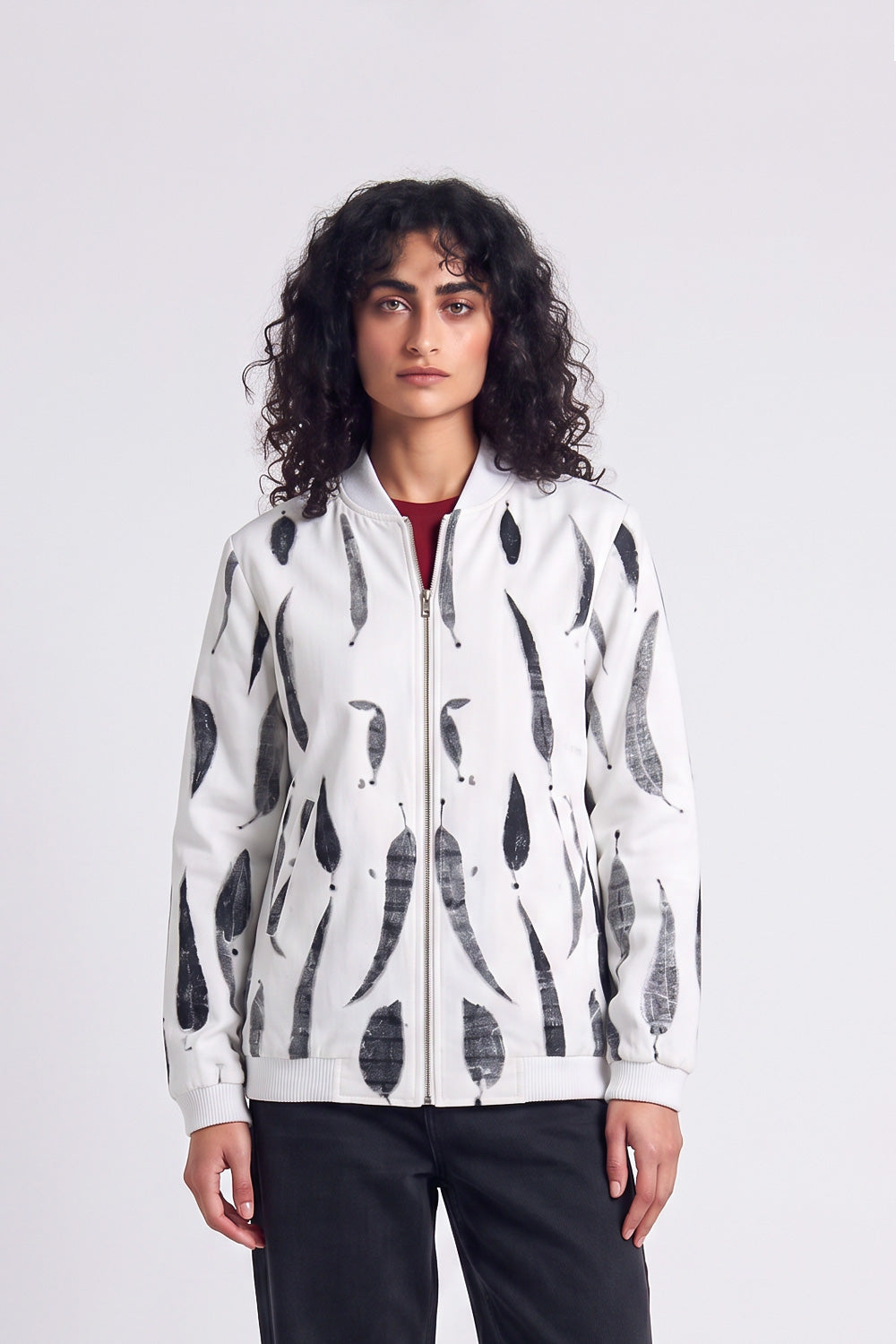 A model with curly dark hair wearing a white zip-up hemp bomber jacket with a black eco-print, styled over a red shirt and black pants, against a light gray studio background.