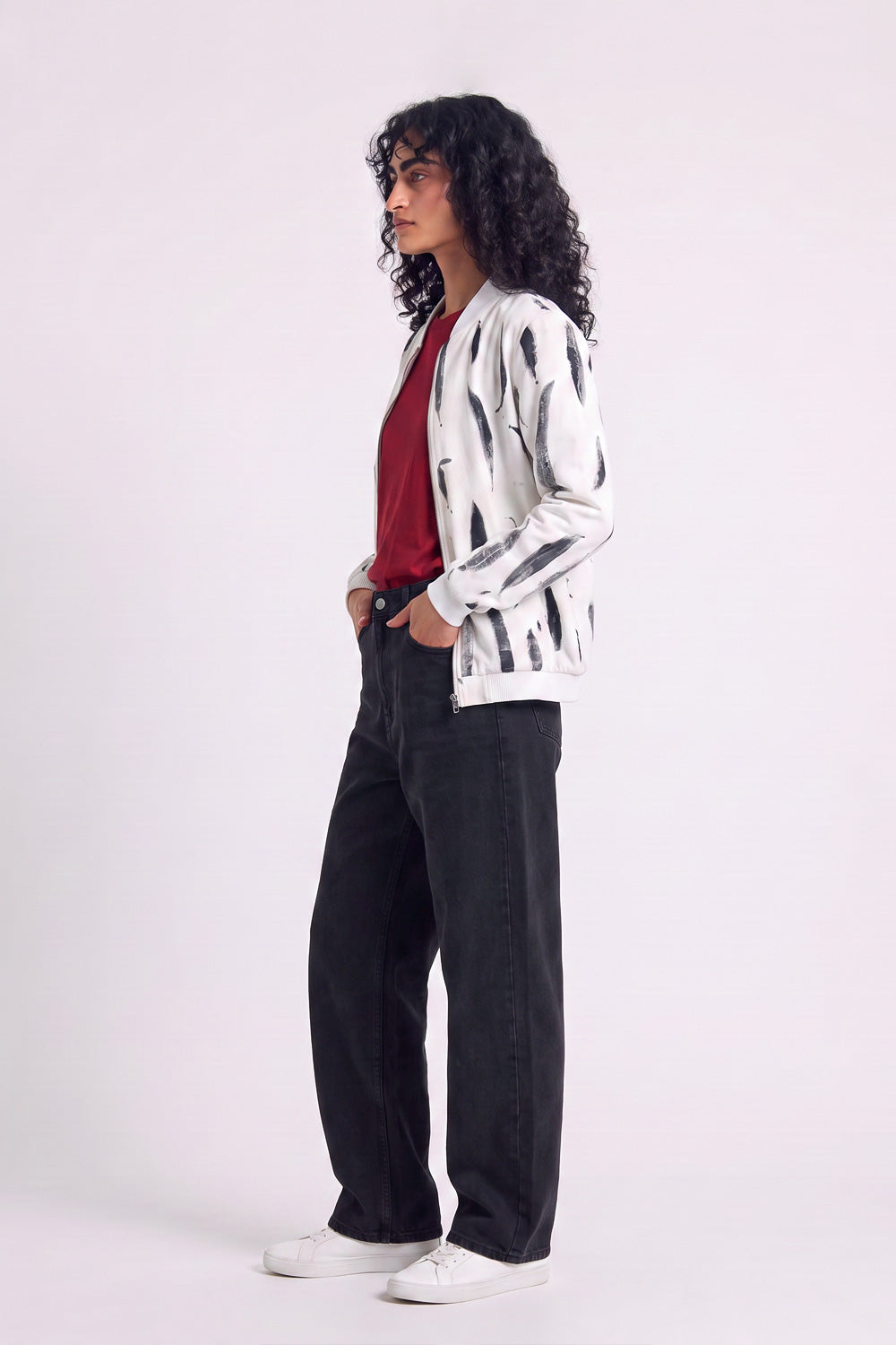 A woman stands in profile wearing a white hemp bomber-style jacket with black botanical print, a red top, black wide-leg jeans, and white sneakers against a plain light background.