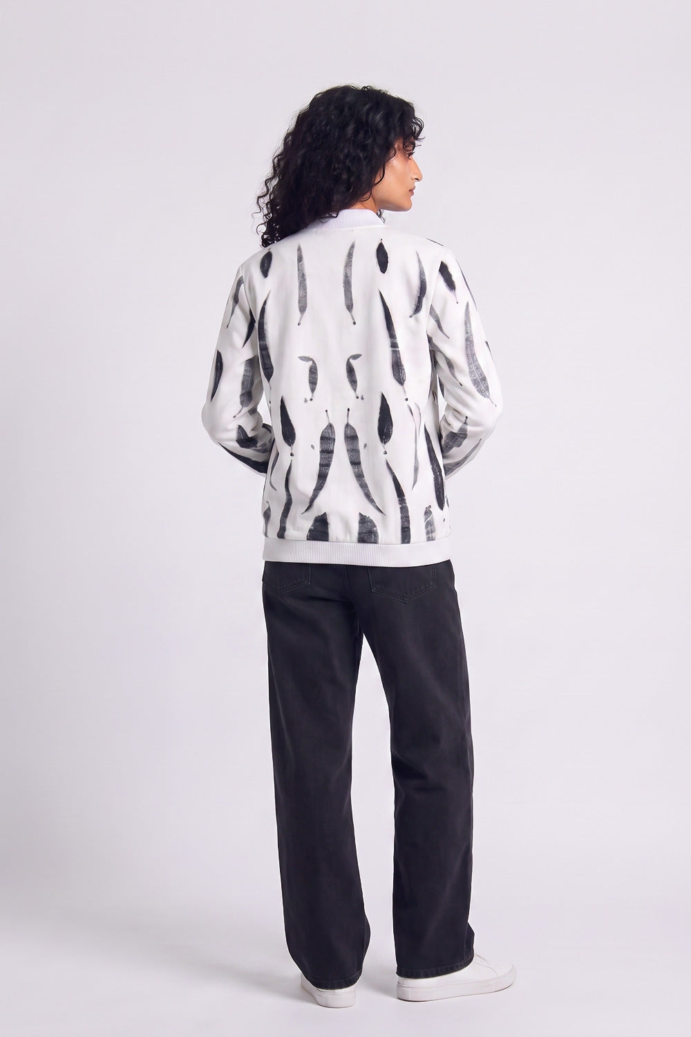 A model shown from the back wearing a white hemp bomber jacket with a black abstract eco-print, paired with black pants and white sneakers, against a light gray studio background.