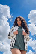 A woman with long curly brown hair and glasses stands outdoors against a bright blue sky with white clouds, wearing a white bomber jacket with black eco-print over a muted green t-shirt dress.