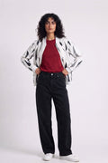 A model stands against a light gray studio background wearing a white hemp bomber jacket with black abstract spots, a red t-shirt, black wide-leg jeans, and white sneakers.