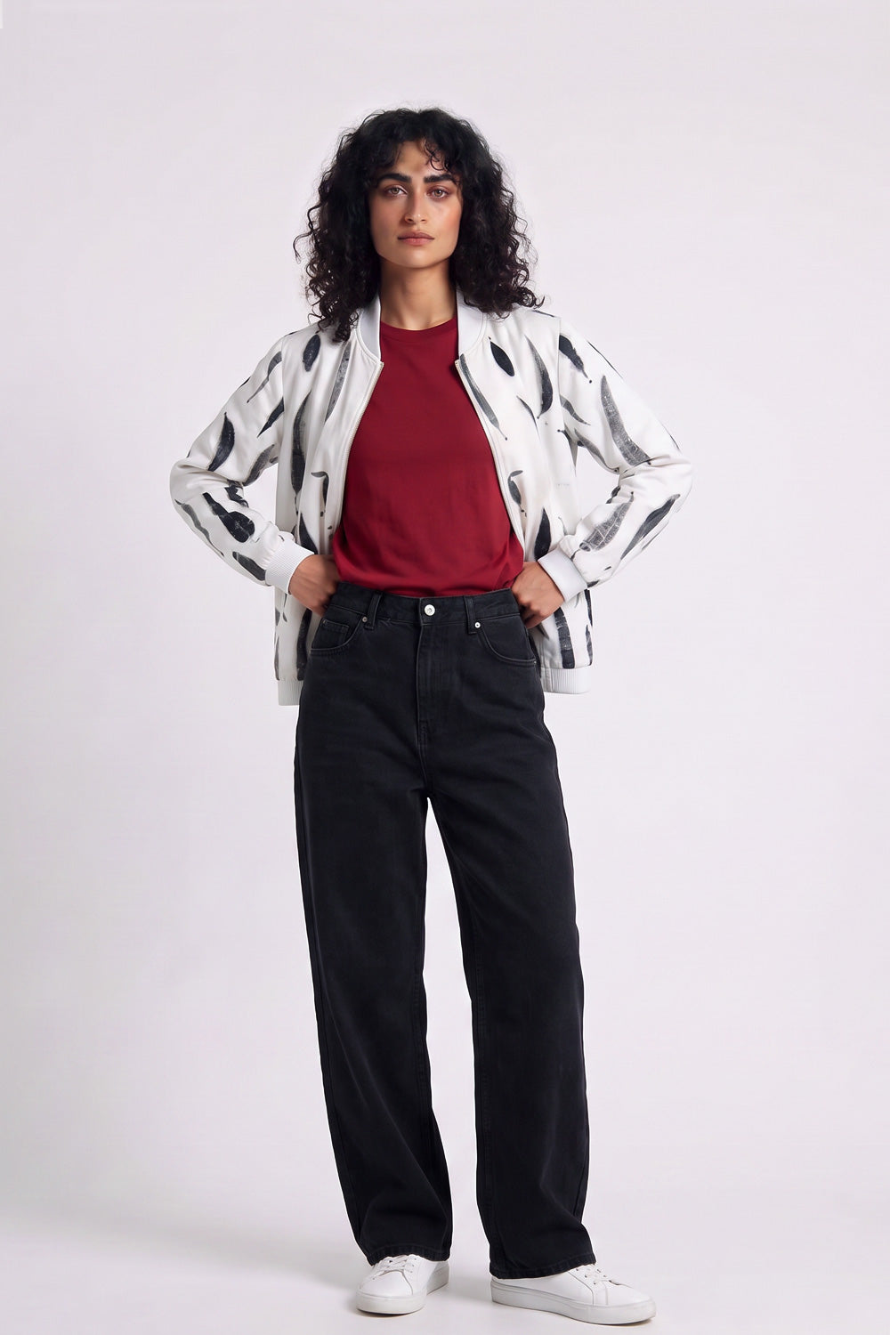 A model stands against a light gray studio background wearing a white hemp bomber jacket with black abstract spots, a red t-shirt, black wide-leg jeans, and white sneakers.