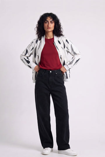 A model stands against a light gray studio background wearing a white hemp bomber jacket with black abstract spots, a red t-shirt, black wide-leg jeans, and white sneakers.