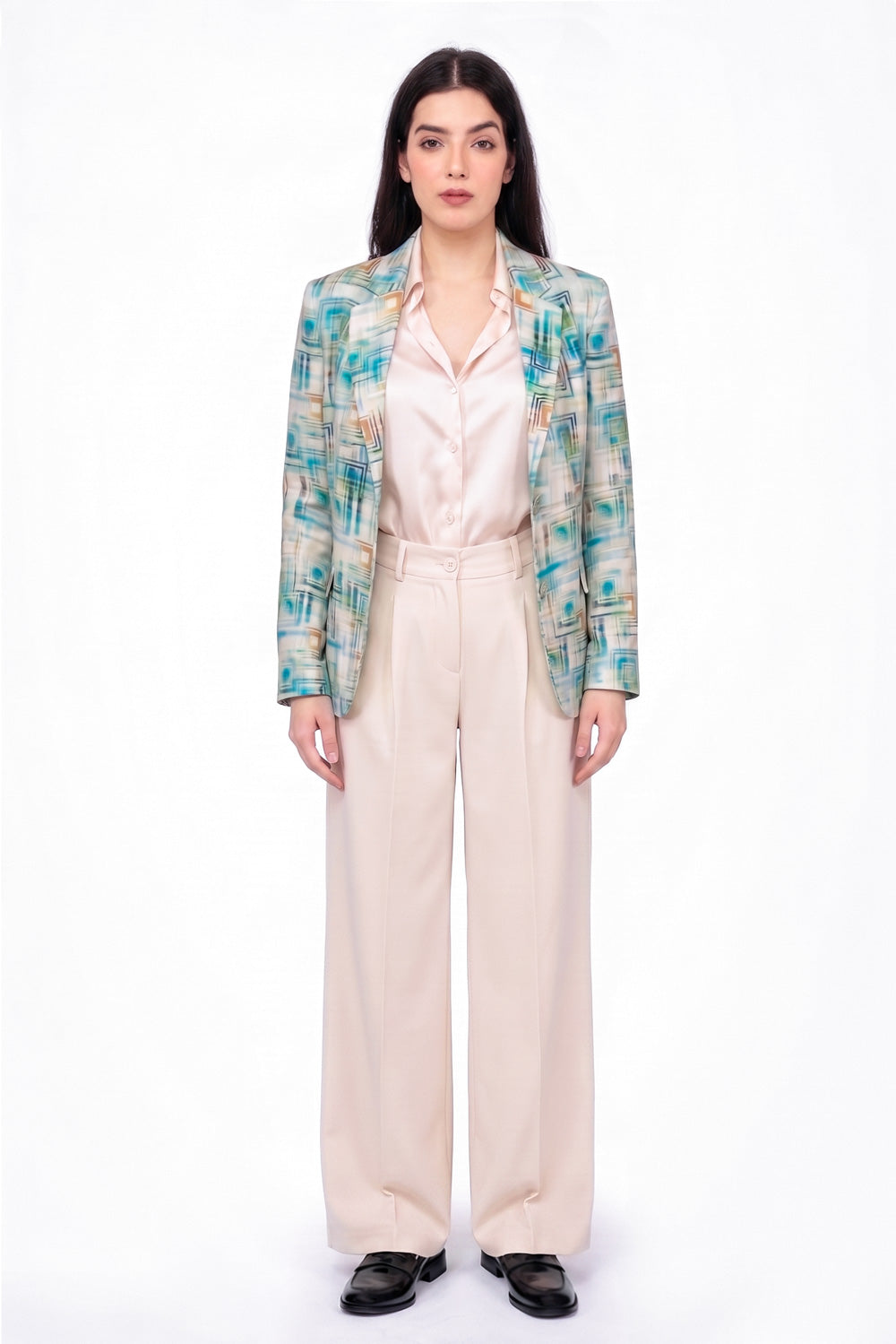 A model stands against a white studio background wearing a long-sleeve patterned linen blazer with teal, green, and beige tones over a pale pink satin shirt and cream wide-leg trousers.