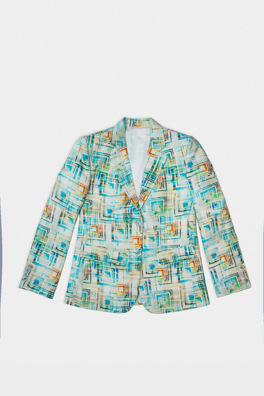 A model stands against a white studio background wearing a long-sleeve patterned linen blazer with teal, green, and beige tones over a pale pink satin shirt and cream wide-leg trousers.