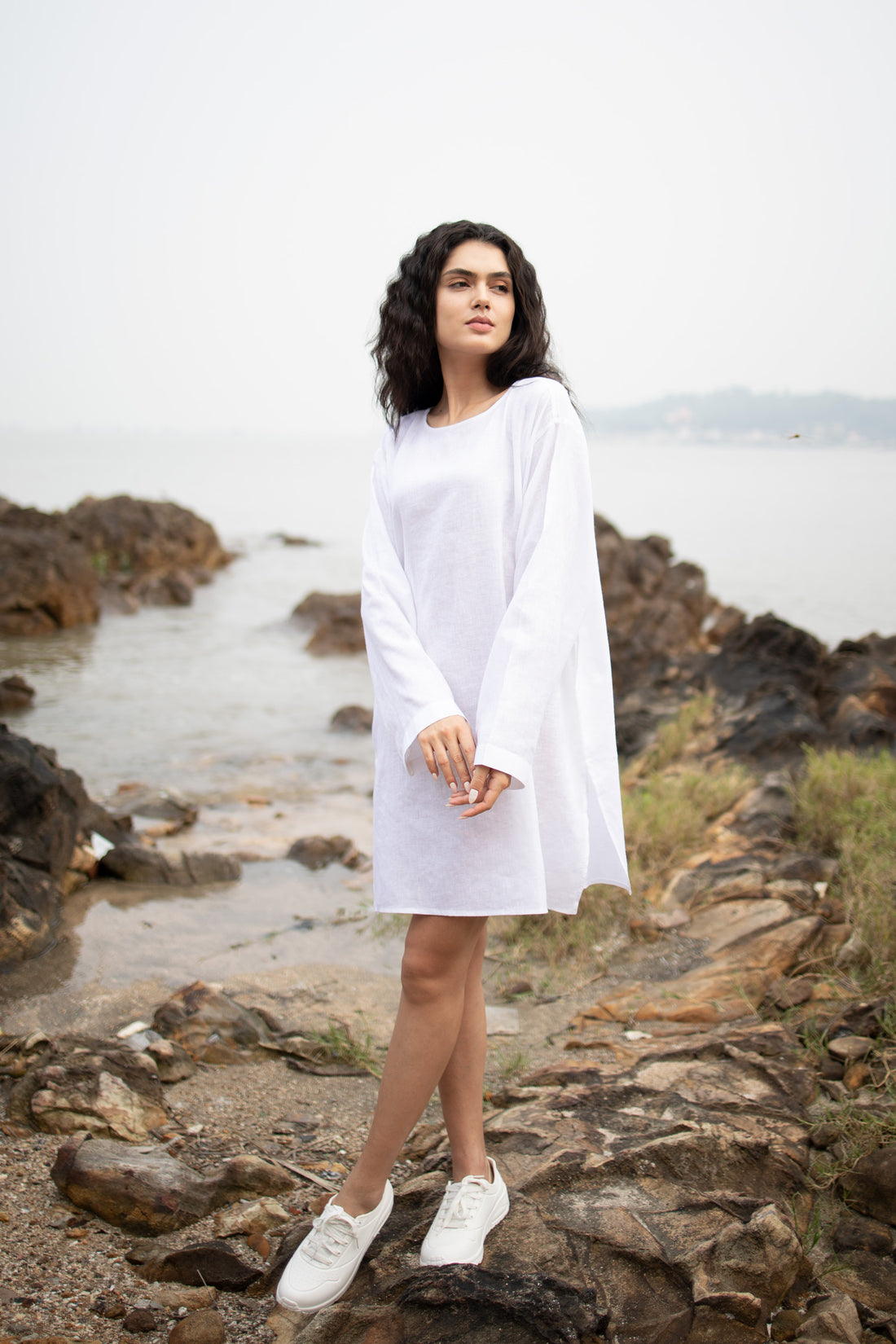 A woman stands on a rocky shoreline wearing a loose white long-sleeve lien tunic dress and white sneakers, with water and distant land visible in the background.