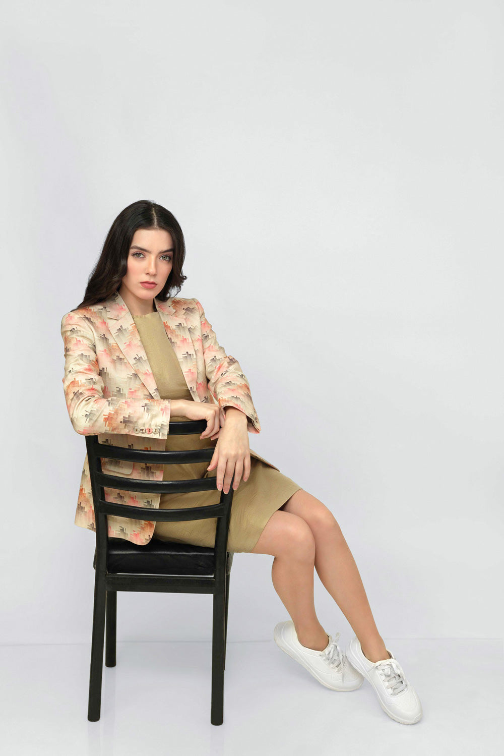 A woman sits on a black wooden chair against a light gray studio background, wearing a beige and pink floral linen blazer over a tan dress with white sneakers.