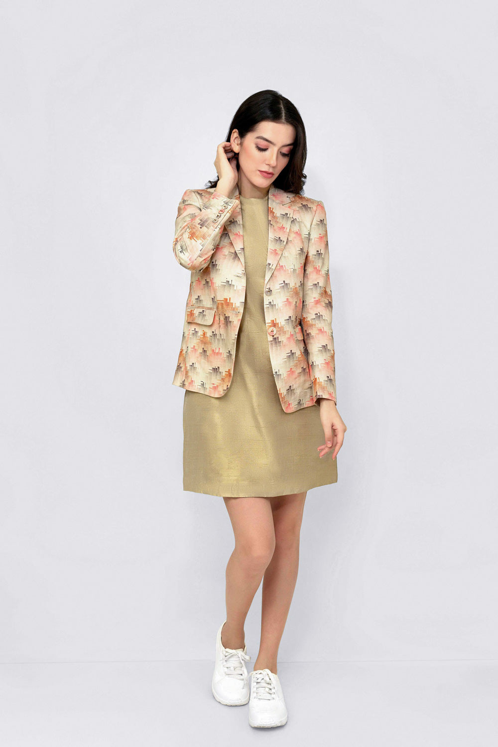 A woman modeling a beige floral linen blazer over a metallic gold midi dress, paired with white sneakers, against a light gray studio background.