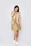 A woman modeling a beige floral linen blazer over a metallic gold midi dress, paired with white sneakers, against a light gray studio background.