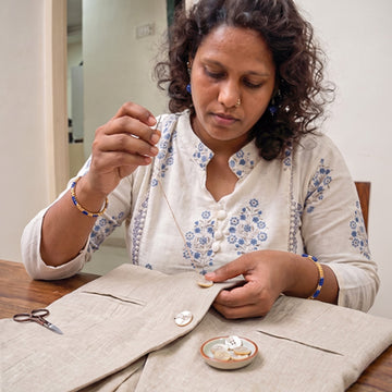 A woman with curly dark hair sits at a wooden table, carefully sewing or buttoning a light beige linen jacket. She is wearing a matching cream-colored embroidered kurta with blue floral patterns.
