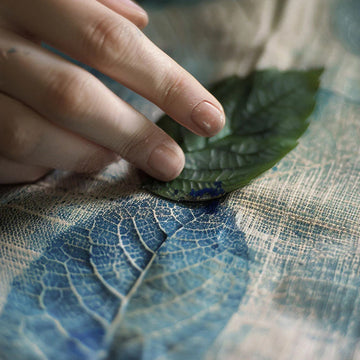 A close-up of a hand resting on a blue and beige patterned fabric surface with a green leaf placed on top.