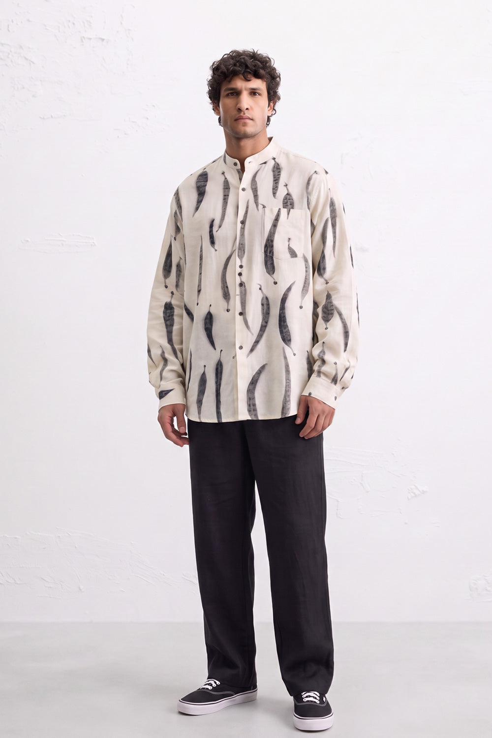 A man modeling a cream-colored long-sleeve button-up shirt with a black abstract leaves eco-print pattern, paired with black trousers and black sneakers, standing against a white textured wall.