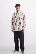 A man modeling a cream-colored long-sleeve button-up shirt with a black abstract leaves eco-print pattern, paired with black trousers and black sneakers, standing against a white textured wall.