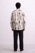 A man viewed from behind wearing a cream-colored long-sleeve button-up shirt with a black abstract leaves eco-print, paired with black trousers and white sneakers, standing against a white textured wall.