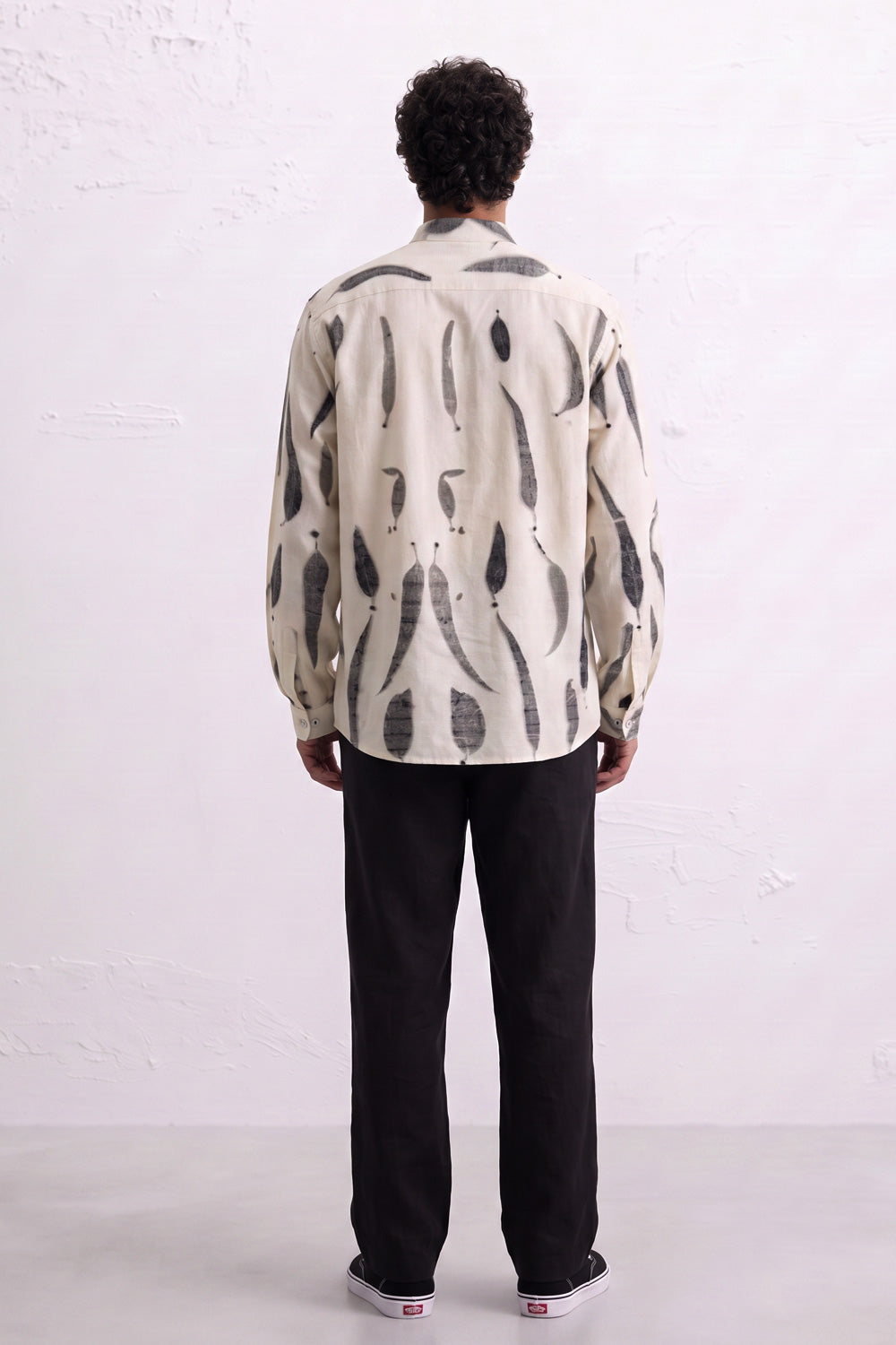 A man viewed from behind wearing a cream-colored long-sleeve button-up shirt with a black abstract leaves eco-print, paired with black trousers and white sneakers, standing against a white textured wall.