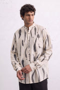 A man wearing a cream-colored long-sleeve button-up shirt with a black leaves eco-print, standing against a white textured wall.