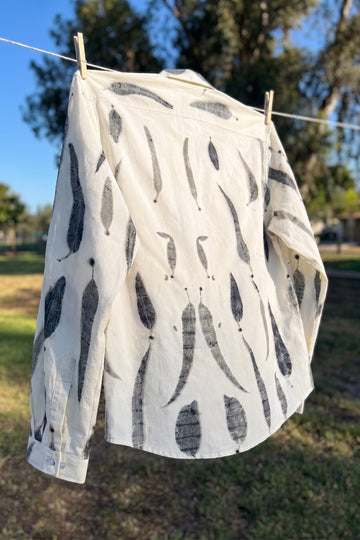 A white long-sleeve shirt with a black leaf eco-print hangs from a clothesline outdoors in a sunny yard with trees and grass in the background.