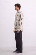 A man wearing a cream-colored long-sleeve button-up shirt with a black abstract leaves eco-print, paired with black trousers and black sneakers, standing in profile against a white textured wall.