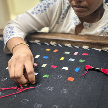A close-up of a person's hand and lower face as they work on a dark fabric panel with colorful embroidered squares and red tassels, wearing a white embroidered blouse.