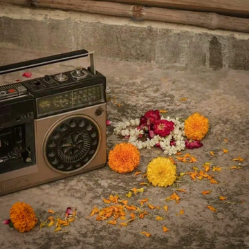A vintage-style boombox radio sits on a concrete floor beside scattered orange marigold petals and flower garlands, with additional marigolds and a small bouquet placed nearby.