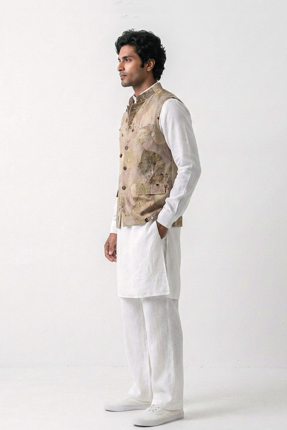 A man modeling a traditional Indian outfit consisting of a white kurta and trousers paired with a beige eco-printed hemp waistcoat, standing in profile against a white studio background.