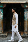 A man in a traditional Indian outfit consisting of a white kurta and trousers with a beige eco-printed hemp vest, walking past a rustic, weathered doorway.